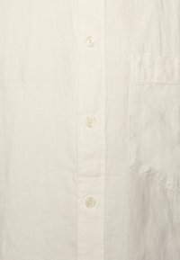 Camisa blanca de botón con tejido ligero, que presenta un bolsillo en el lado izquierdo del pecho, con botones de plástico transparente y una superficie texturizada.