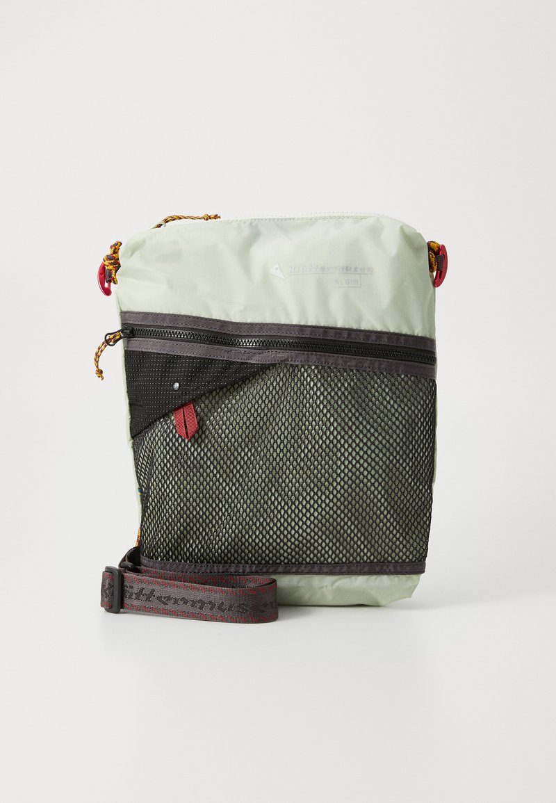 Light green crossbody bag with a black mesh front pocket, adjustable strap, and red accents. Features zippers and a textured surface.