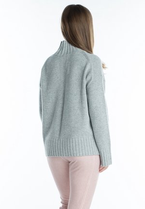 Femme portant un pull col roulé côtelé gris clair et un pantalon rose pâle, debout de dos sur un fond blanc.
