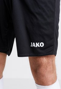 Juodos sportinės šortai, pagaminti iš lengvos, lygaus audinio. Kairėje apačioje yra baltas logotipas "JAKO". Paprastas dizainas be raštų.