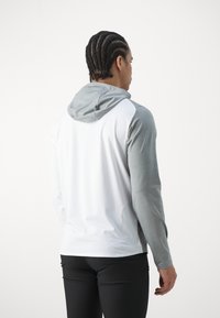 Haut à capuche léger en blanc et gris. Il a une poche à l'avant, une capuche ajustable et des manches longues en matériau et texture contrastants.