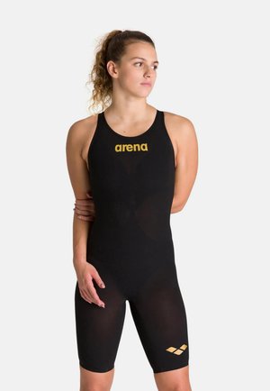 Nuotatrice con costume intero nero con logo "arena" giallo, posa con le braccia dietro la schiena su sfondo bianco.