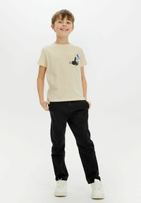 Camiseta de algodón beige con un diseño impreso en negro de un cartón de leche derramando líquido negro, combinada con pantalones negros y zapatillas blancas.