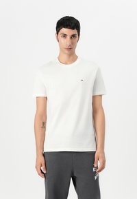 T-shirt en coton de couleur claire avec un col rond, des manches courtes et un petit logo sur la poitrine. Porté avec un pantalon de survêtement gris foncé.
