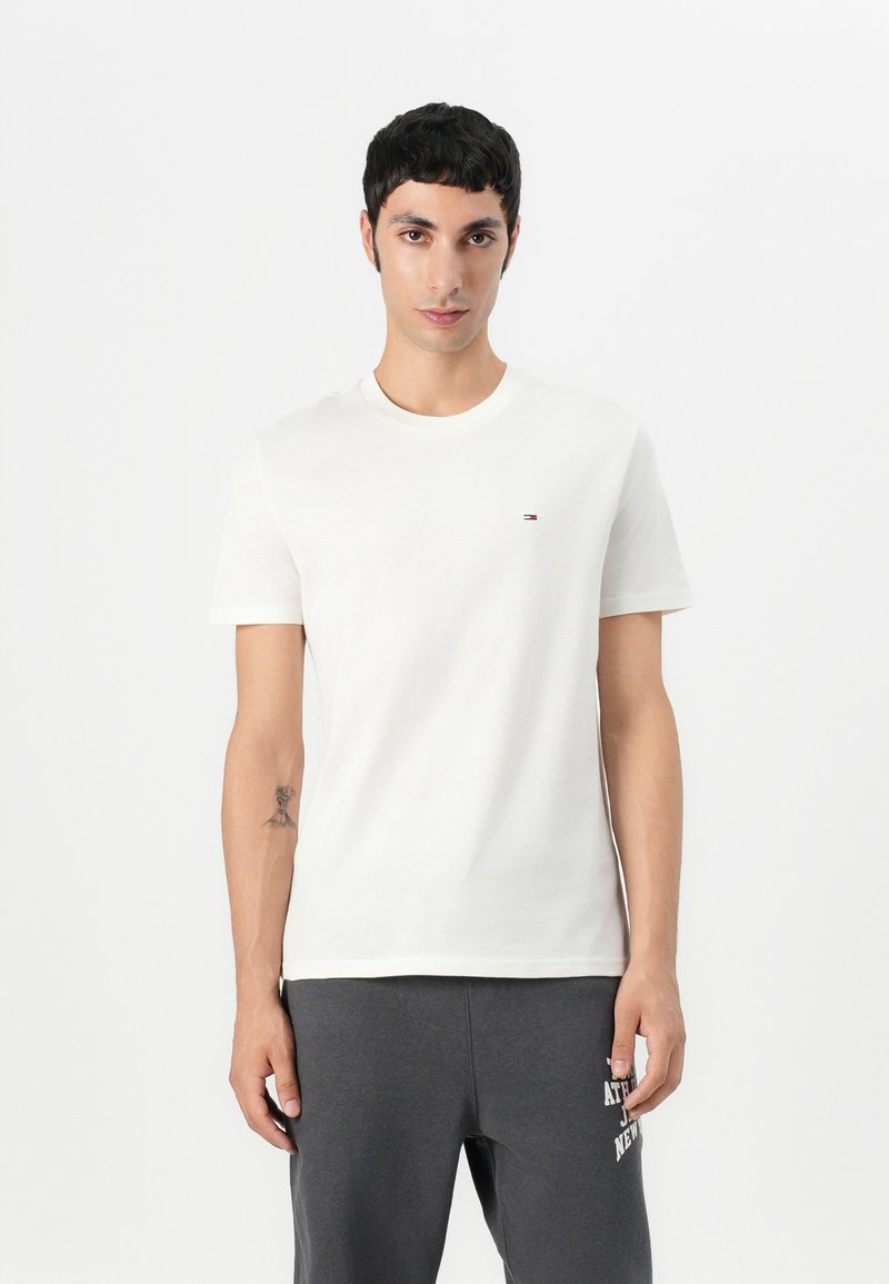 Camiseta de algodón de color claro con cuello redondo, mangas cortas y un pequeño logo en el pecho. Se lleva con pantalones de chándal gris oscuro.