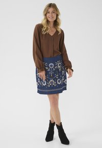 Blouse marron à manches longues avec un col en V, associée à une jupe bleu foncé ornée de broderies florales. Des bottines noires à la cheville complètent la tenue.