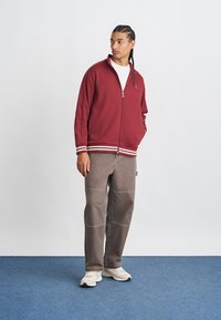 Veste zippée bordeaux avec des rayures blanches aux poignets et à l'ourlet, associée à un pantalon gris avec des coutures visibles et des baskets blanches.