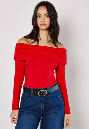 Haut rouge à manches longues, dénudé des épaules, avec un détail froncé. Associe à un jean bleu taille haute et une ceinture noire avec une boucle ronde.