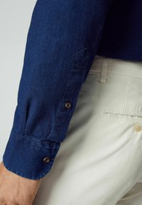 Manica di camicia in denim blu scuro con chiusura a bottone, caratterizzata da un polsino arrotondato e una texture sottile, abbinata a pantaloni beige.