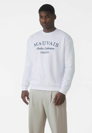 Biely mikina s modrým vyšívaným logom s textom "MAUVAIS Atelier Collection MMXVIII." Má rebrované manžety a lem. Kombinovaná so svetlými nohavicami.