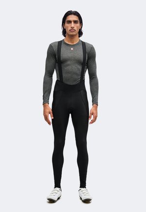 Mann steht und trägt ein eng anliegendes graues Langarmshirt, schwarze Radhose mit Trägern und weiße Radschuhe vor einfachem Hintergrund.