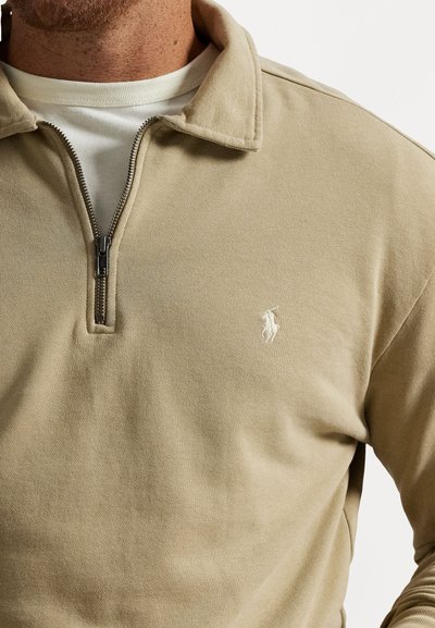 Pull en beige à zip quart porté sur un t-shirt blanc à col rond, avec un petit logo de joueur de polo blanc brodé sur la poitrine gauche.