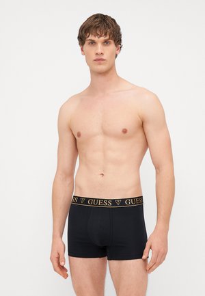 Boxer briefs pretos com um logo dourado "GUESS" na cintura. O tecido é suave e tem um ajuste justo que acentua a silhueta.