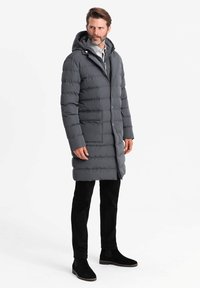 Cappotto lungo grigio imbottito con cappuccio, dotato di zip frontale e bottoni a pressione, tasche laterali e un tessuto liscio e resistente all'acqua.