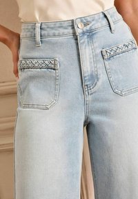 Lichtblauwe spijkerbroek met gevlochten zakaccenten, hoge taille en recht gesneden ontwerp. Stevige textuur met minimale vervagingdetails.