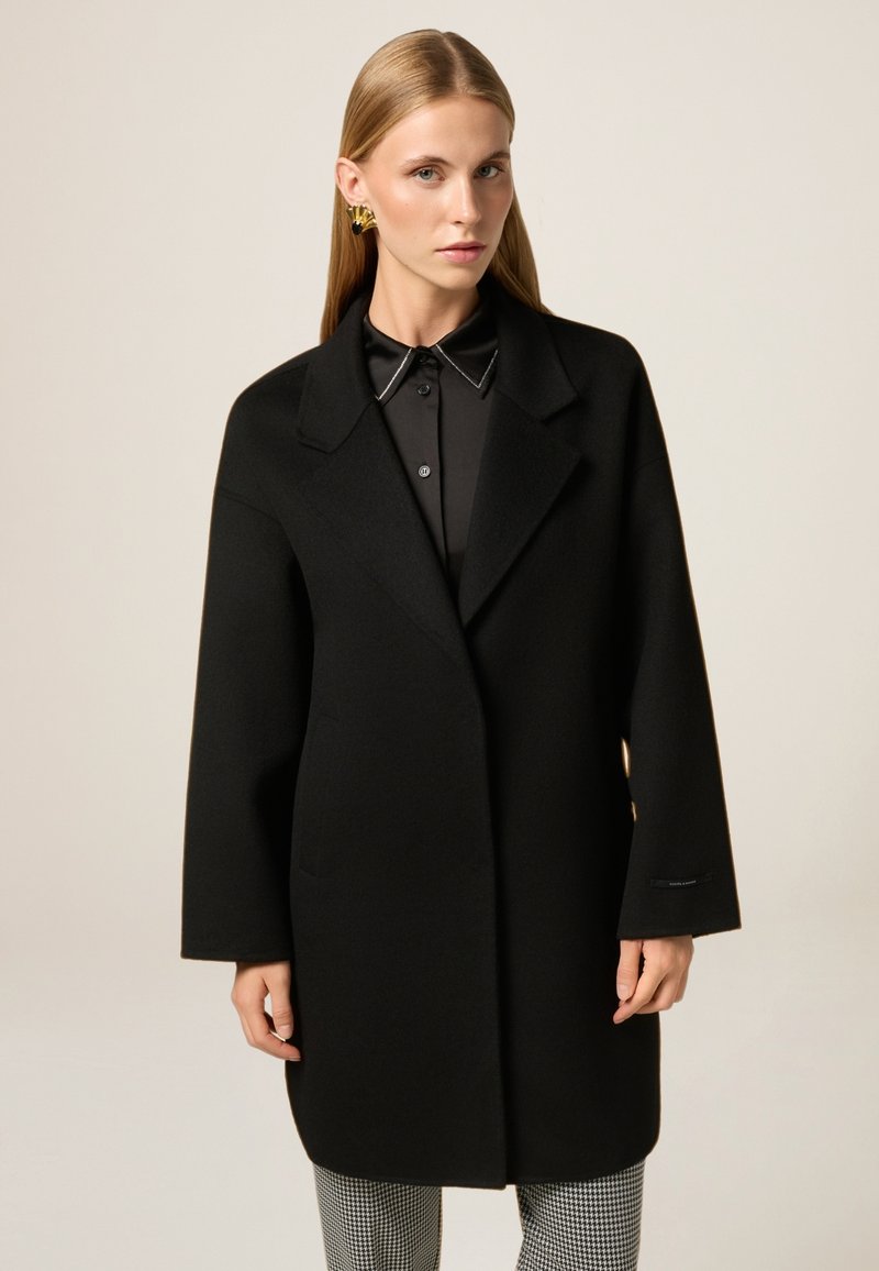 Cappotto di lana nera con un taglio ampio, rever a punta, tasche laterali e maniche lunghe, abbinato a una camicia nera con colletto sotto.