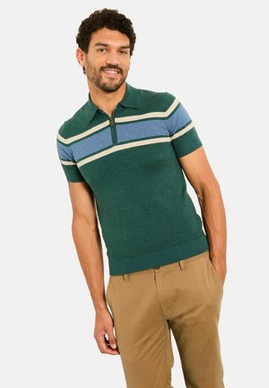 Mann lächelt und lehnt sich leicht, trägt einen grünen kurzärmligen Pullover mit blauen und beigen Streifen sowie beige Hosen, die Hände in den Taschen.