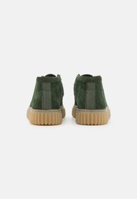 Un paio di stivaletti alla caviglia in suede verde con suole in gomma beige testurizzate, visti da dietro su uno sfondo bianco.