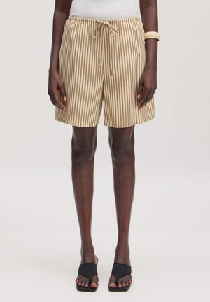 Personne portant un short beige et blanc à rayures verticales avec cordon, un haut blanc, des sandales noires ouvertes à l'avant et un bracelet crème au bras gauche.