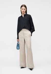 Γυναίκα που φοράει χαλαρό μαύρο πουκάμισο με κουμπιά, μπεζ παντελόνι με ευρύ πόδι, μαύρα μυτερά παπούτσια, κρατώντας ένα μικρό μπλε τσαντάκι.