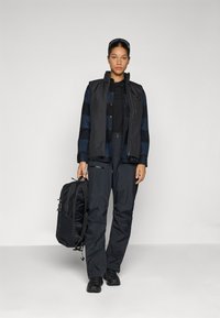 Zwarte vest over een marineblauwe en zwart geruite shirt; zwarte waterdichte broek; houdt een zwarte rugzak vast; strakke design en praktische materialen.