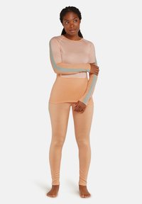Donna che indossa un top a maniche lunghe color pesco e leggings con strisce grigie sulle maniche e ai lati, in piedi scalza su uno sfondo bianco.