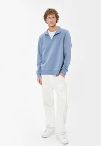 Felpa zip-up di colore azzurro chiaro in tessuto morbido, con colletto e polsini a coste, abbinata a pantaloni cargo bianchi e sneakers bianche.