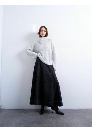 Kvinde iført en grå oversize rullekrave trøje, lang sort nederdel og sorte ankelstøvler, der står mod en hvid væg.