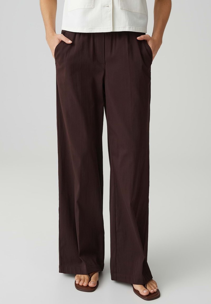 Persoon draagt donkerbruine broek met wijde pijpen, handen in de zakken en bruine sandalen met vierkante neus, tegen een effen achtergrond.