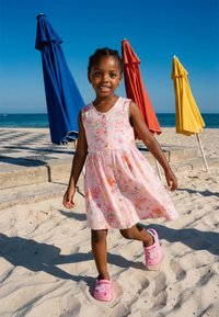 Jeune fille en robe fleurie rose et chaussures roses marchant sur une plage de sable avec des parasols fermés bleus, rouges et jaunes sous un ciel clair et l'océan.