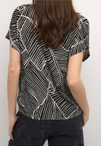 Frau mit langem braunem Haar, trägt ein kurzärmliges Oberteil mit abstrakten schwarz-weißen Streifen und dunkle Jeans, mit dem Rücken zur Kamera gewandt.