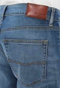 Τζιν παντελόνια σε μεσαίο μπλε χρώμα, με πίσω τσέπη, ορατή ραφή σε ανοιχτό καφέ και καφέ δερμάτινη ετικέτα της μάρκας στη μέση.