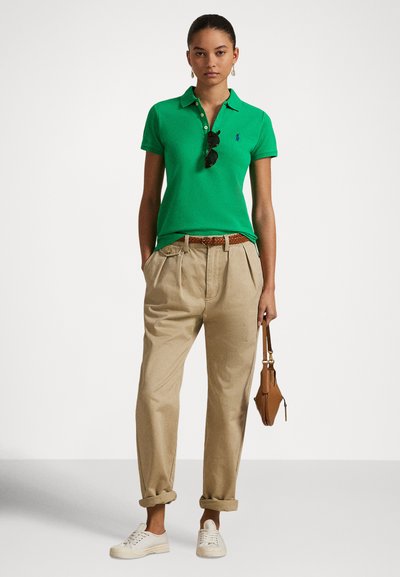 Grønn pikéskjorte med krage, beige vide bukser, hvite joggesko og en liten brun håndveske. Solbriller henger fra skjorten.