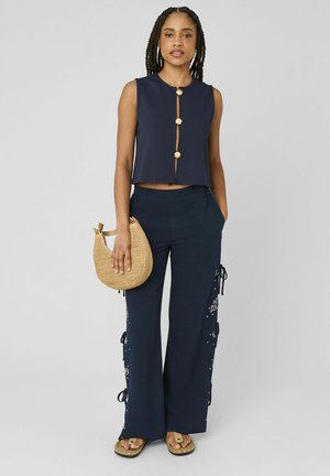 Femme portant un haut sans manches bleu marine avec des boutons dorés, un pantalon large bleu marine avec des détails sur les côtés, tenant un sac à main rond tissé et portant des sandales.