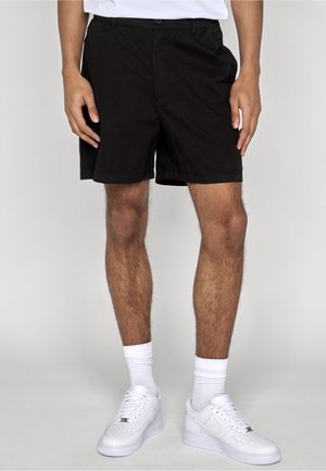 Mand iført sorte shorts, hvide sokker og hvide sneakers, stående mod en neutral baggrund.