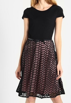 Vrouw die een zwart shirt met korte mouwen draagt en een roze rok tot op de knie met een zwarte geruite mesh-overlay en een dunne zwarte taille riem.