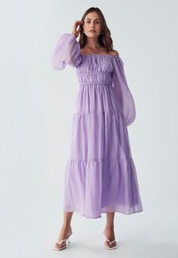 Violetti olkapäiltä avattu maksimekko kevyestä kankaasta, jonka vyötärö on joustava, kerroksellinen muotoilu, pitkät puff-hihat ja viehättävä siluetti.
