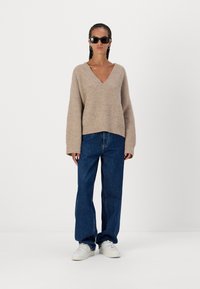 Pulover bej din tricot cu decolteu în V, manți lungi și croială lejeră, asortat cu blugi albaștri cu talie înaltă și adidași albi.