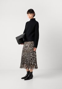 Vrouw in een zwarte kabelgebreide coltrui, met een geplooide midi-rok met luipaardprint, zwarte enkellaarsjes, en met een grote zwarte clutch in de hand.