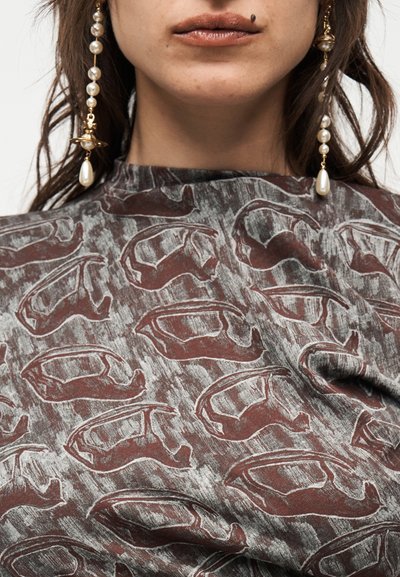 Κοντινό πλάνο του κάτω μέρους του προσώπου και του λαιμού ενός ατόμου που φοράει ένα καφέ και γκρι τοπ με σχέδια αφηρημένων σχημάτων και μακριά σκουλαρίκια με μαργαριτάρια.