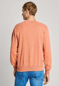 Sudadera naranja claro con mangas largas y puños acanalados, con cuello redondo y un corte relajado, combinada con unos vaqueros azules.