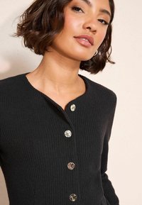 Crni rebrasti kardigan s okruglim dekolteom, s pet velikih prozirnih dugmadi duž prednje strane za zatvaranje.