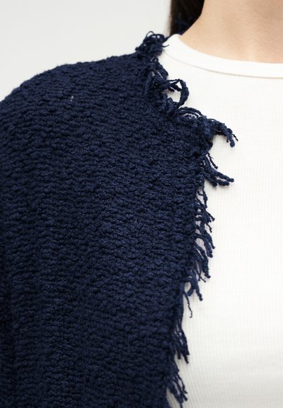 Κοντινό πλάνο ενός ατόμου που φοράει μπλε σκούρο, υφασμένο, με κρόσσια πλεκτή ζακέτα πάνω από ένα λευκό ριμπ κορμάκι.