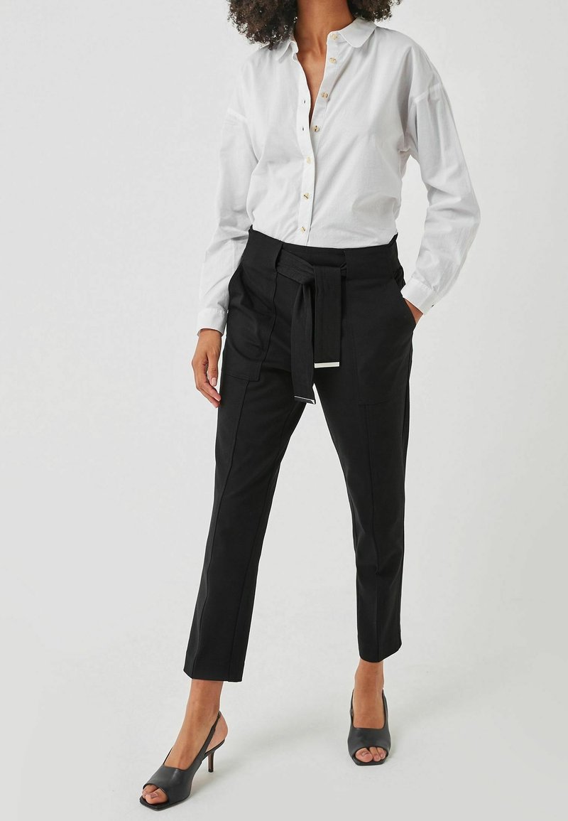 Chemise blanche boutonnée avec col, boutons dorés et coupe ample, associée à un pantalon noir taille haute avec ceinture à nouer et poches.