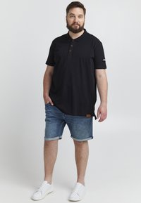 Sort polo-shirt med brune knapper, parret med blå denimshorts og hvide sneakers. Poloen har et logo på den ene ærme og en struktureret overflade.