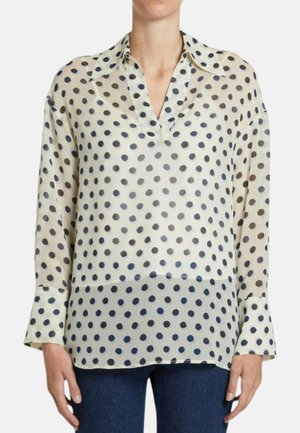 Donna che indossa una camicetta color crema a maniche lunghe con pois scuri e colletto, abbinata a jeans blu scuro, in piedi su sfondo bianco.