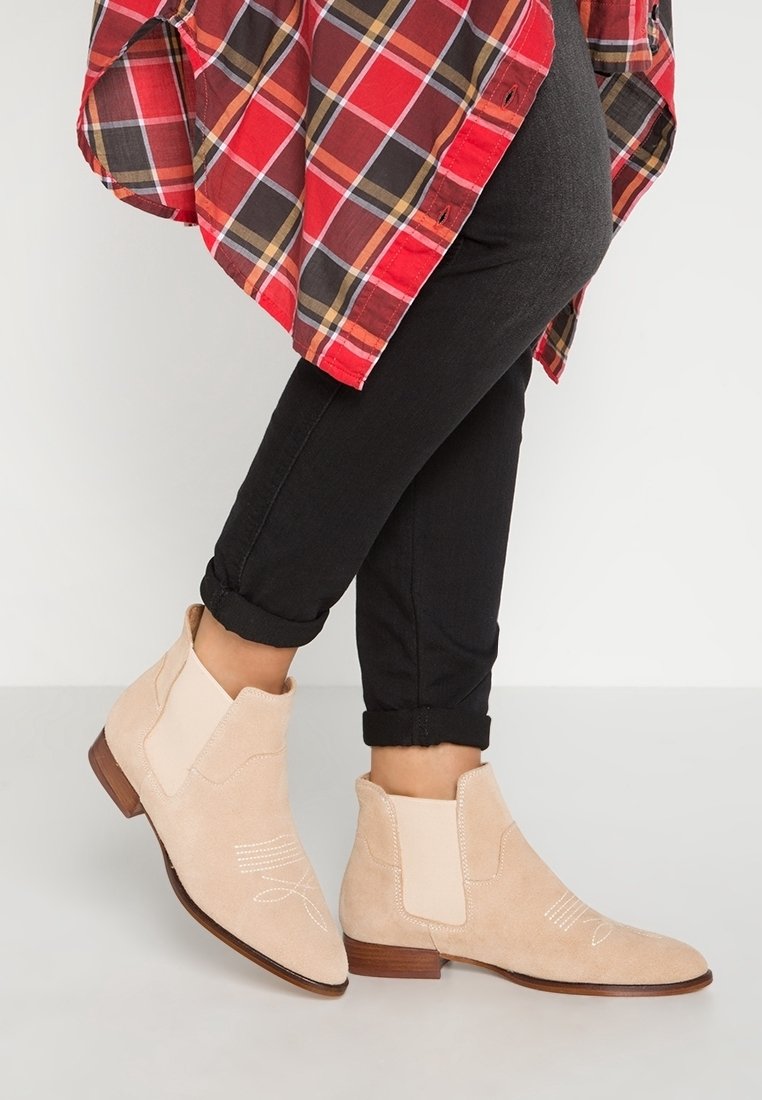 Bottines en daim beige à détails de couture, portées avec un pantalon noir retroussé et une chemise à carreaux rouge.