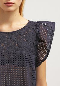 Blouse bleu marine avec une partie supérieure en dentelle florale texturée et une partie inférieure en mesh à motifs. Comprend des manches courtes à volants et un col rond.