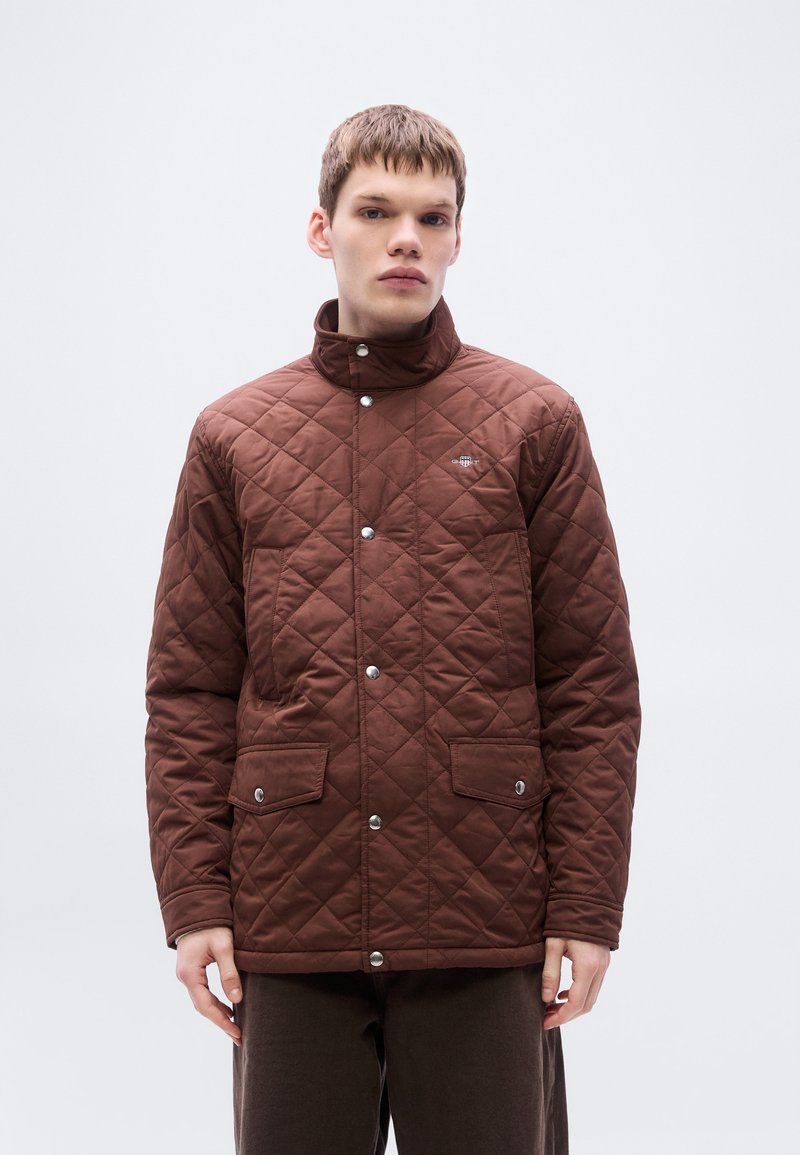 Veste matelassée marron avec un col montant, dotée de deux poches avant et de boutons-pression. Texture lisse avec un motif en losanges.