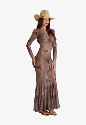 Donna che indossa un lungo vestito marrone con fantasia, cappello di paglia a tesa larga e sandali, in piedi con una mano sul fianco.